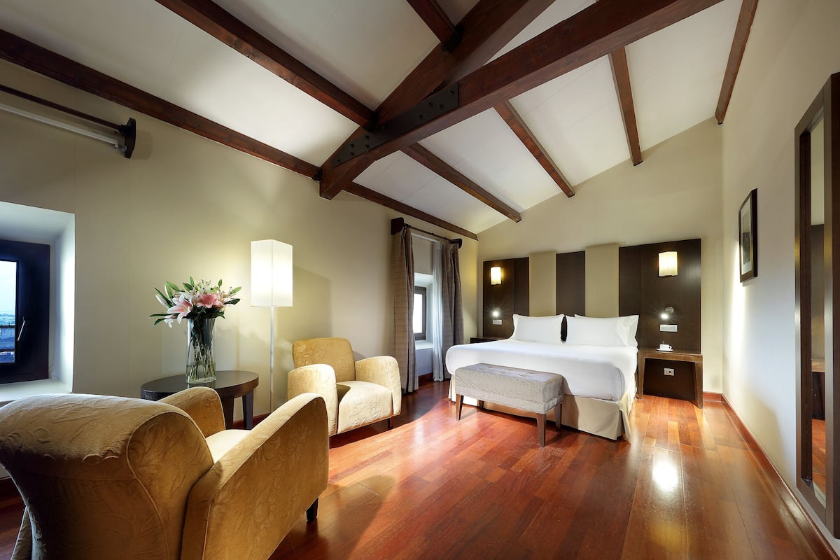 A hotel room with a wooden ceiling, a large bed, two armchairs, a small table with a vase of flowers, and a nightstand with a lamp.