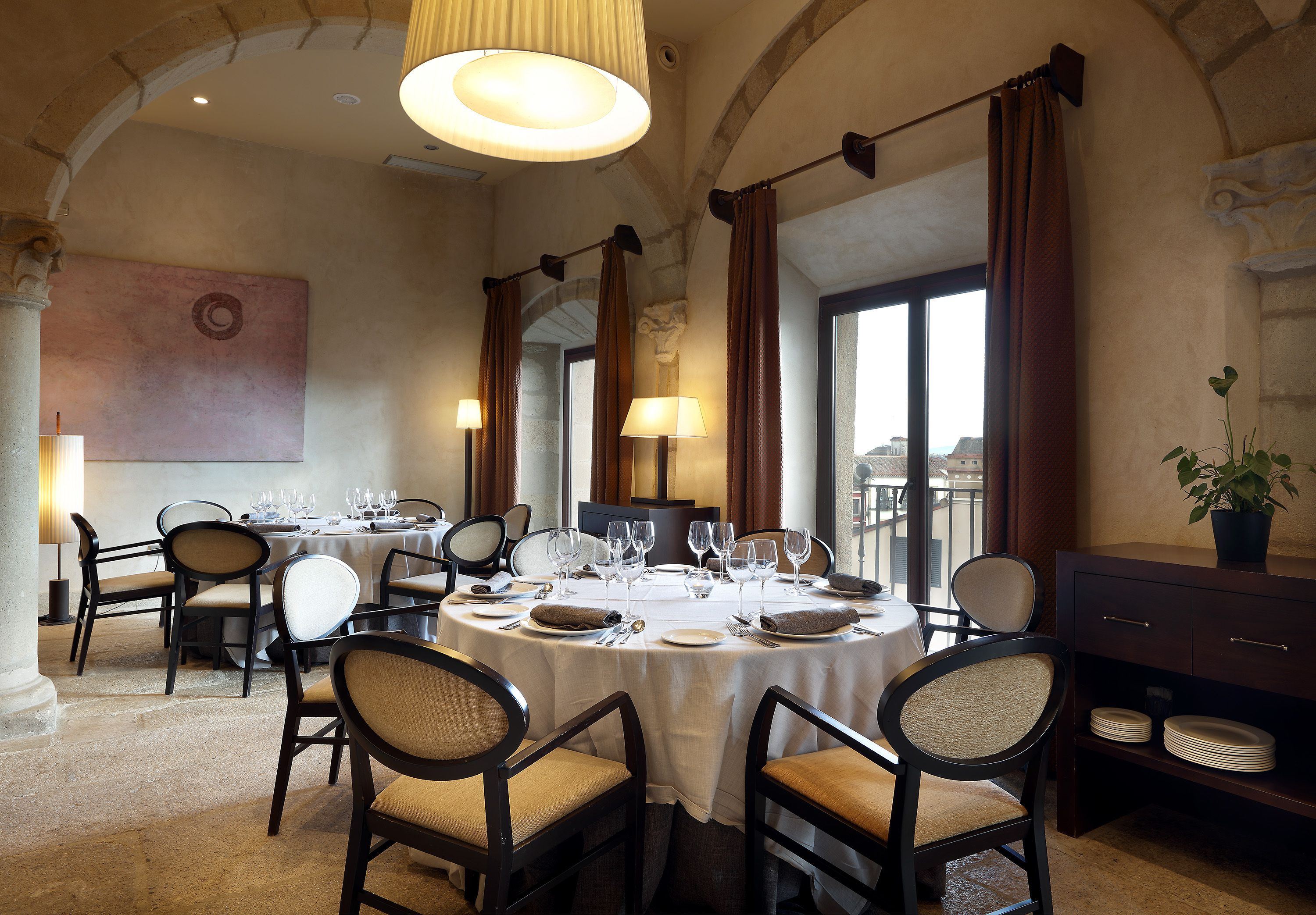 Un comedor con mesas redondas dispuestas para una comida, sillas y vistas de la ciudad a través de amplias ventanas.