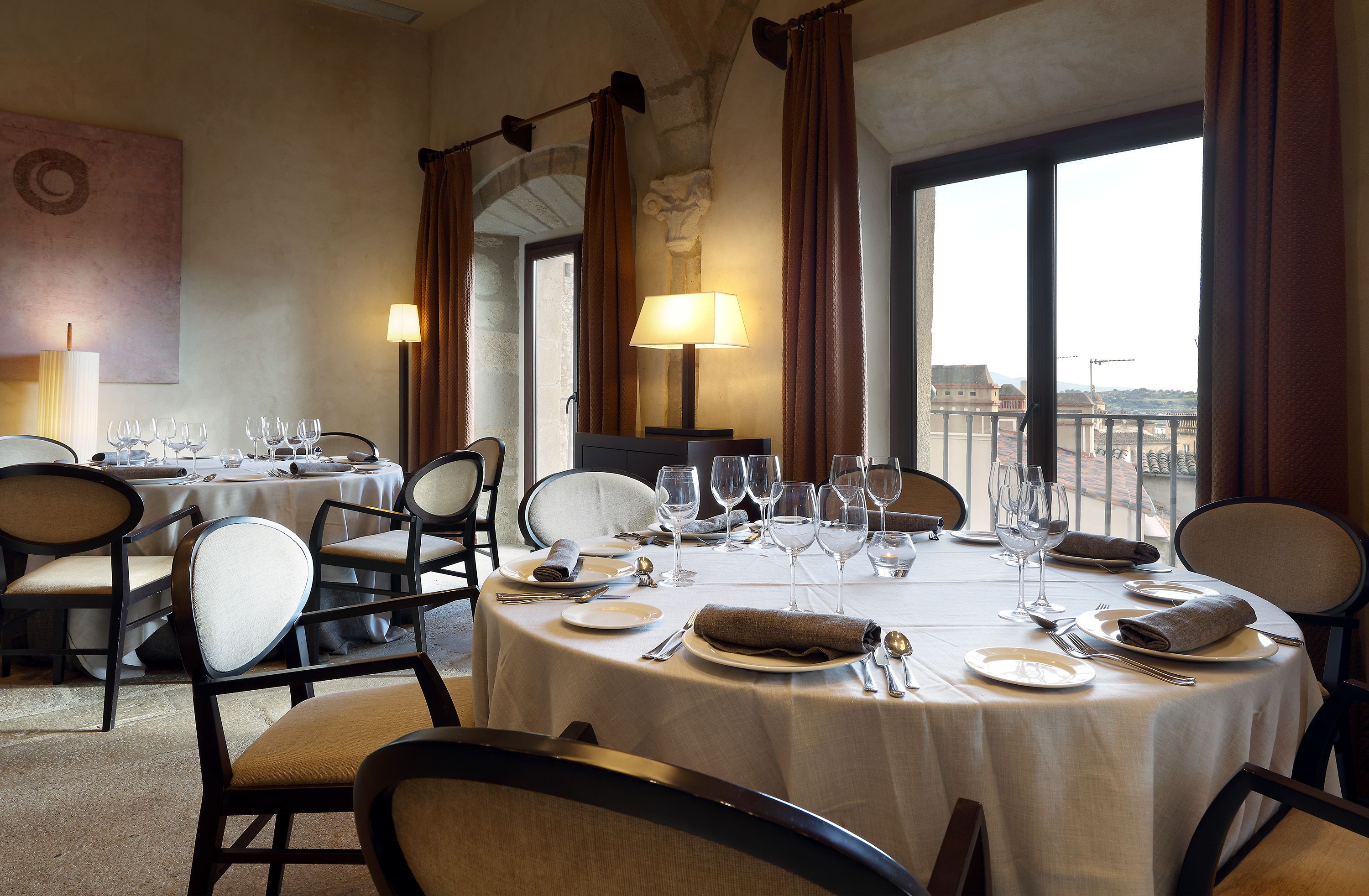 Un comedor con mesas redondas dispuestas para una comida, sillas y vistas de la ciudad a través de amplias ventanas.