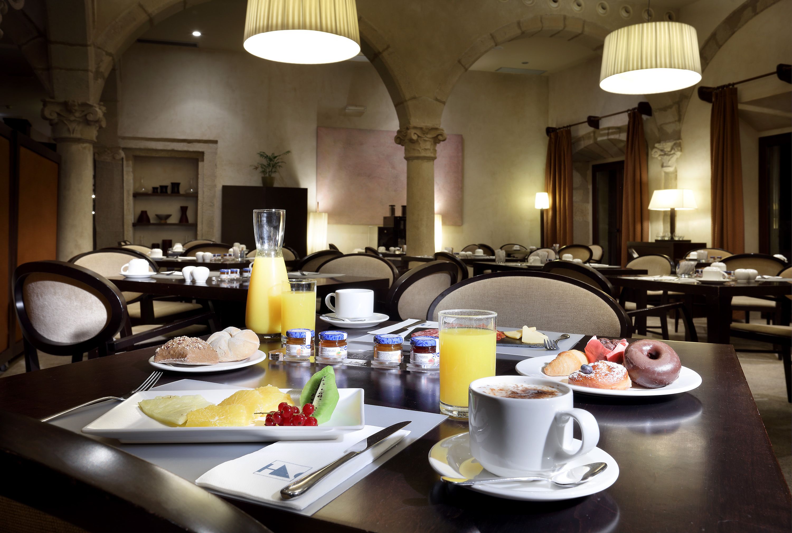 A breakfast setting with a variety of dishes, including pastries, fruits, and beverages, in a dining area with multiple tables and chairs.