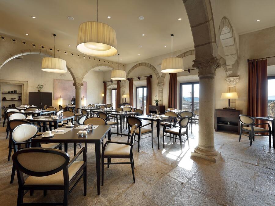 A spacious dining area with multiple tables and chairs, large hanging lamps, and a view of the city through large windows.