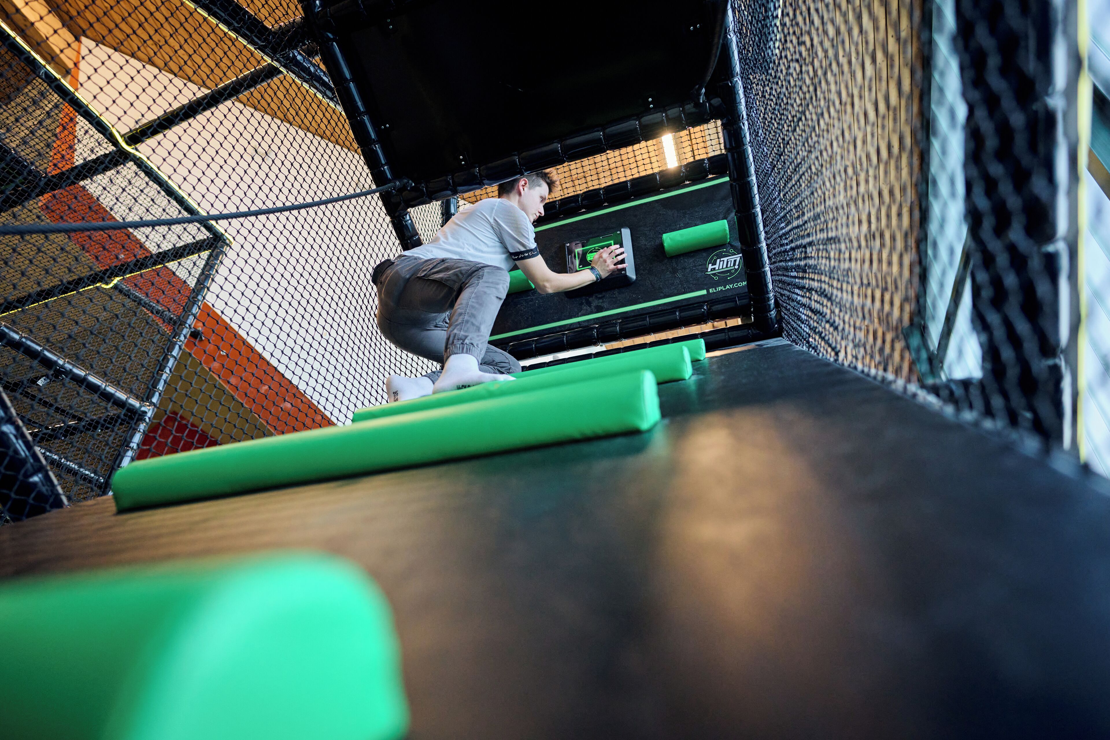 A person climbing a netted obstacle course with green mats and a black wall.