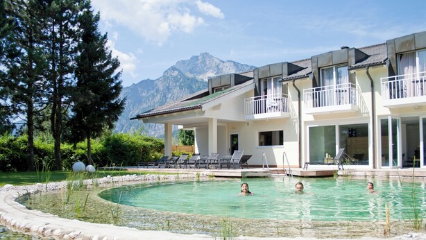 Una piscina con agua cristalina, rodeada por un borde de piedrecitas, frente a un edificio con balcones y un paisaje de montañas.