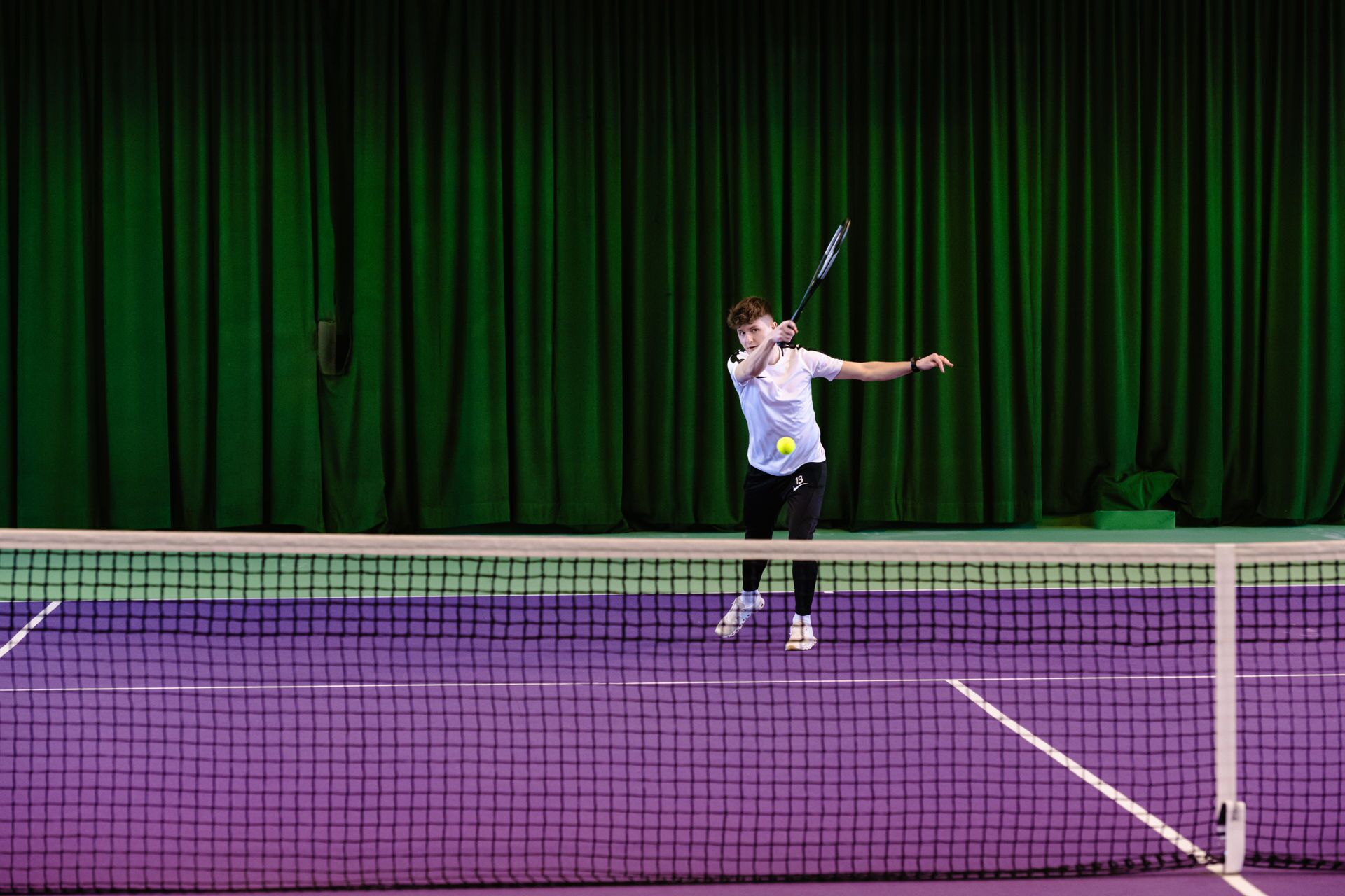 A tennis player in action on a purple court with a green curtain backdrop.