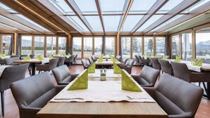 Un restaurante con una larga mesa dispuesta para una comida, rodeado de sillas y con vistas a montañas a través de amplios ventanales.