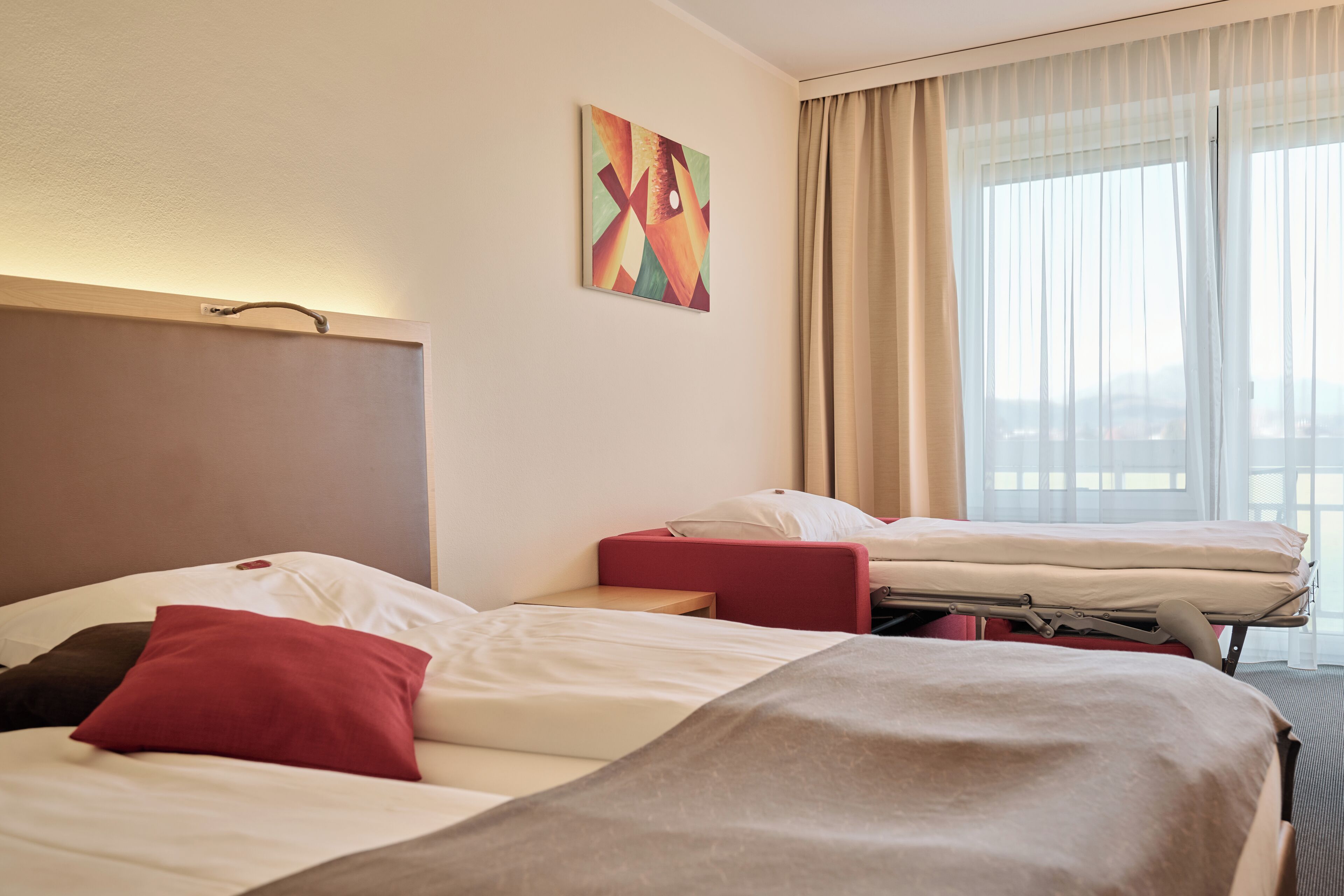 Habitación de hotel con dos camas, un cojín decorativo rojo, un cabecero color beige y vista a la ciudad desde la ventana.