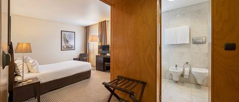 A hotel room with a bed, bedside table, lamp, and a wooden bench.