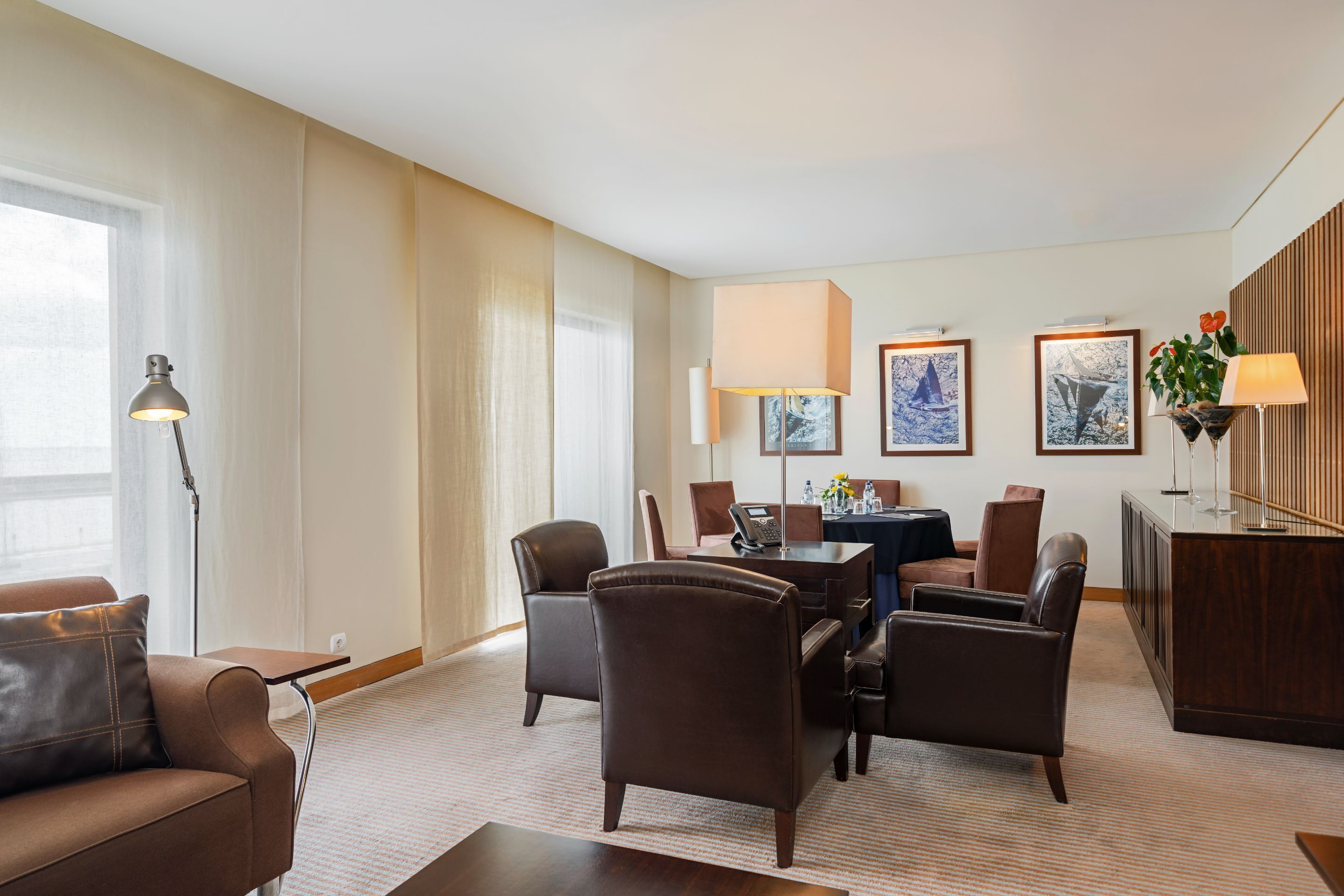 Ein Hotelzimmer mit Sofa, Stühlen, Schreibtisch und Esstisch. An den Wänden hängen Bilder und auf der Kommode steht eine Pflanze.