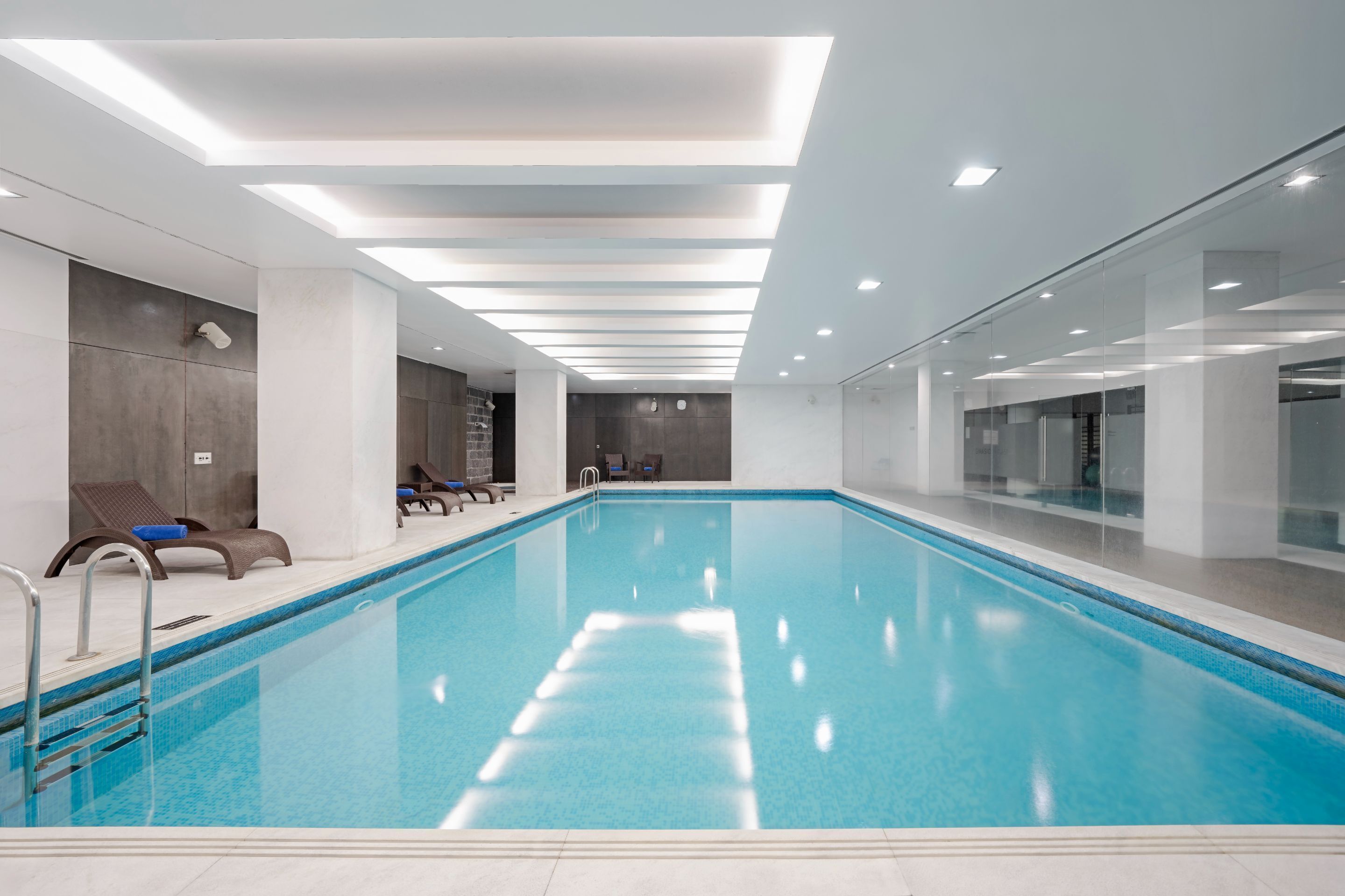 An indoor swimming pool with clear blue water, surrounded by modern lounge chairs and a glass wall.