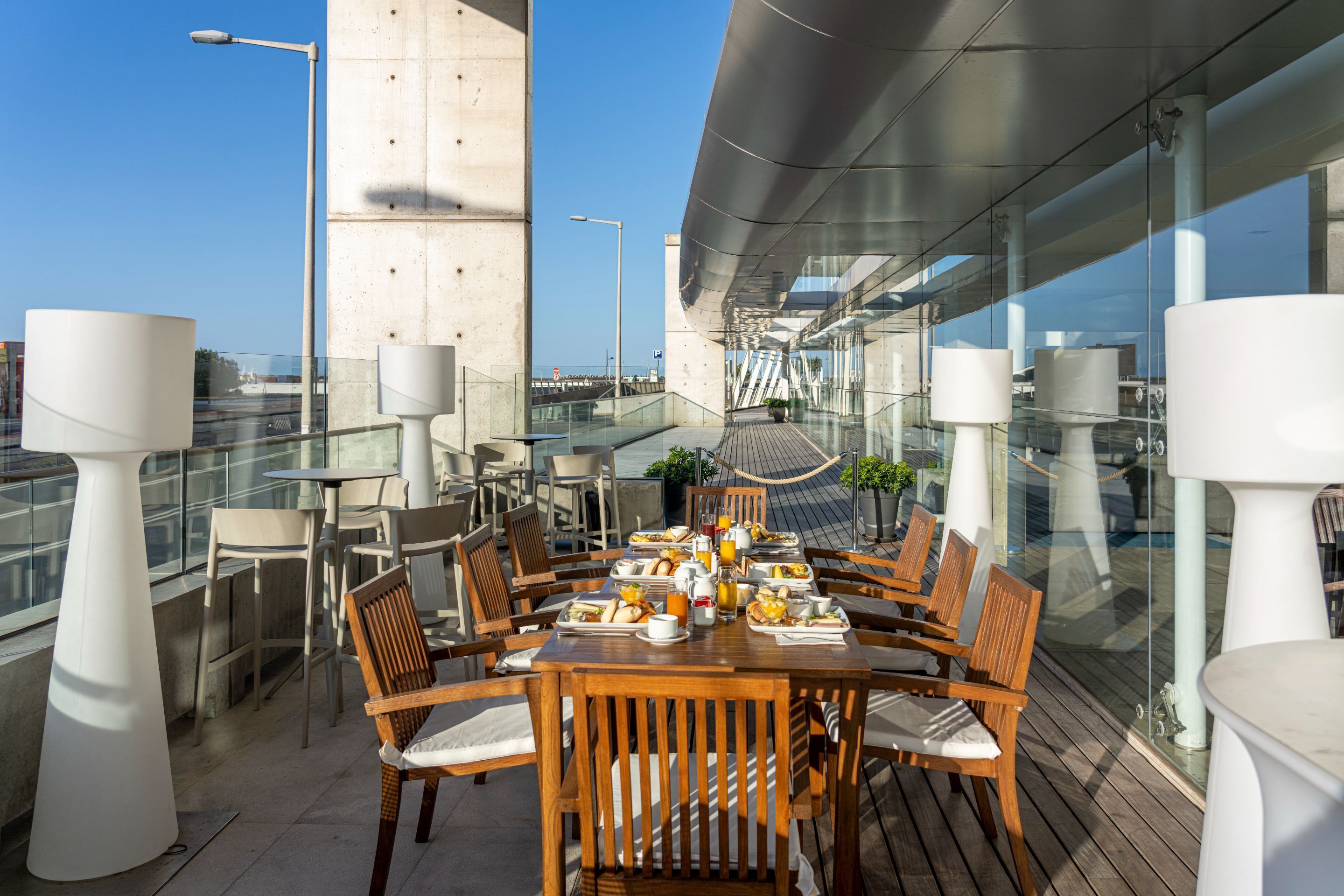 An outdoor dining area with wooden tables and chairs, a variety of dishes and drinks, and a modern architectural backdrop.