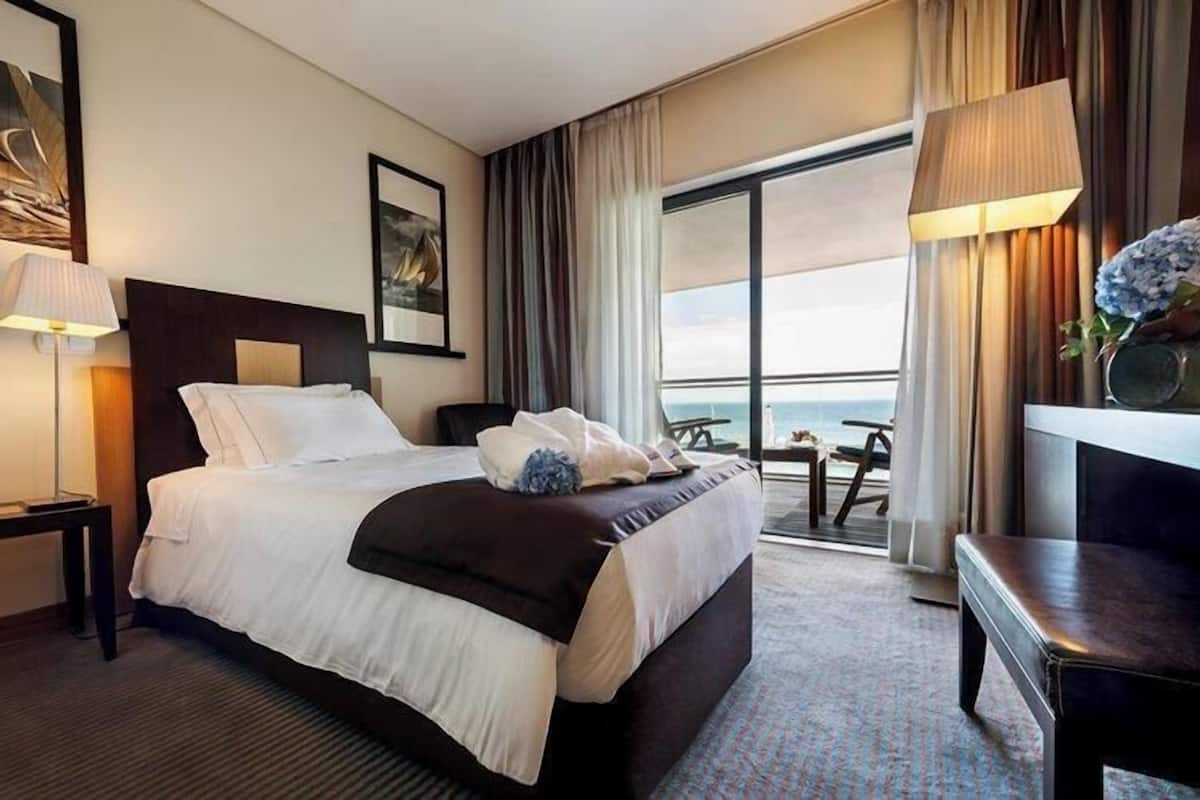Ein Hotelzimmer mit Bett, Schreibtisch, Stuhl, Lampe, einem Gemälde und Blick auf das Meer.