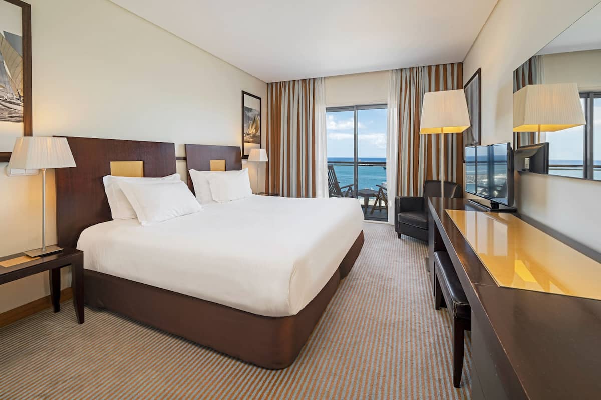 Ein Hotelzimmer mit einem großen Bett, einem Schreibtisch, einem Sessel, einem Fernseher und Blick aufs Meer.