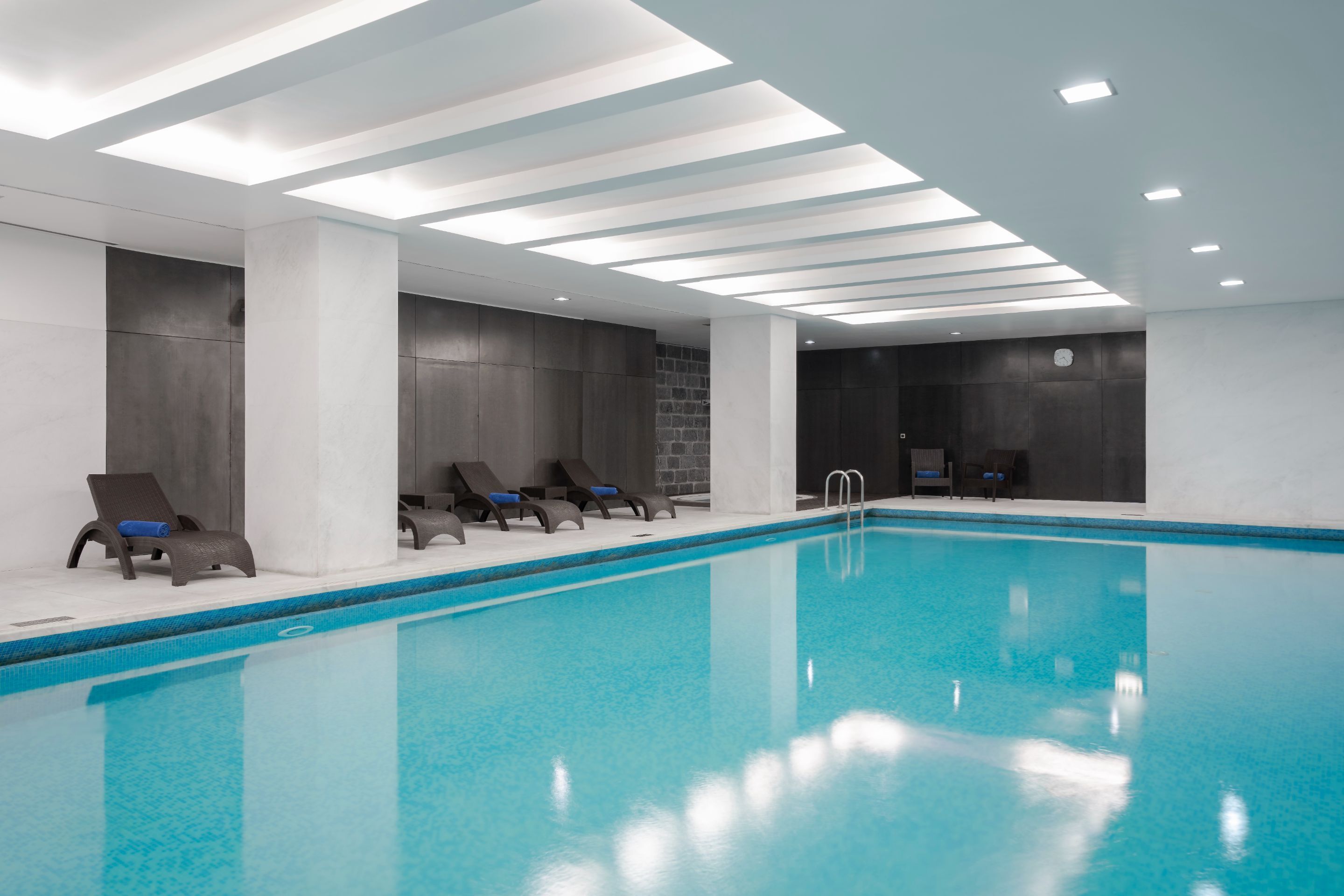An indoor swimming pool with clear blue water, surrounded by lounge chairs and a modern ceiling design.