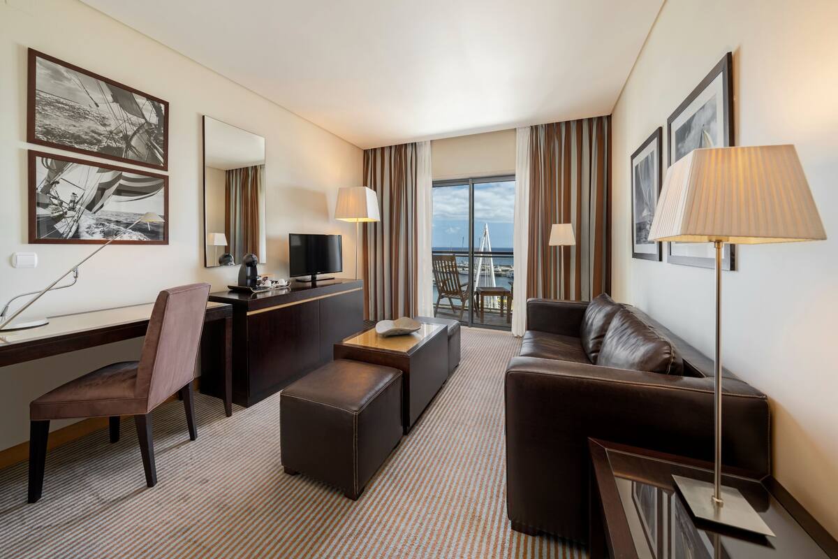Ein Hotelzimmer mit großem Fenster, Schreibtisch mit Stuhl, einer Couch und einem Fernseher.