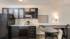 A compact kitchen with a refrigerator, microwave, and dishwasher. There's a small dining table with a chair and a wall-mounted lamp. - Candlewood Suites Washington Dulles Sterling by IHG (Sterling)