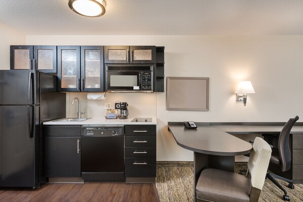 A compact kitchen with a refrigerator, microwave, and dishwasher. There's a small dining table with a chair and a wall-mounted lamp. - Candlewood Suites Washington Dulles Sterling by IHG (Sterling)