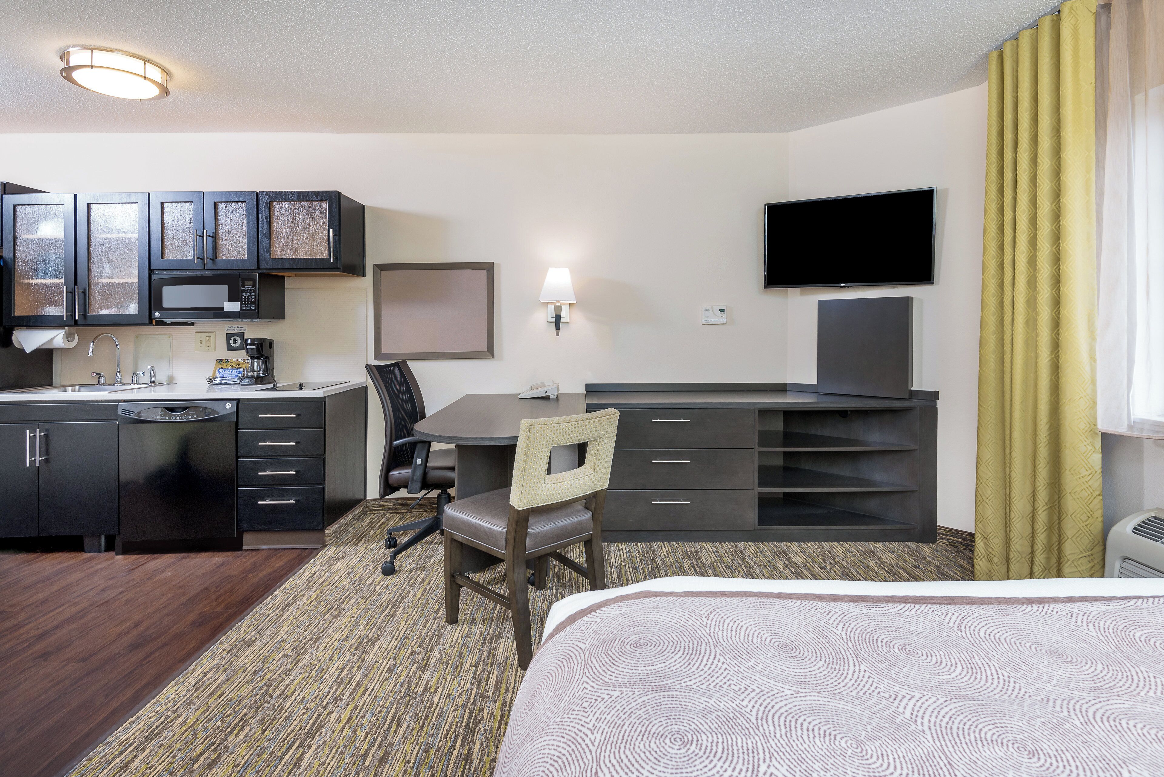 A hotel room with a bed, a desk with a chair, a microwave, a coffee maker, a TV, and a window with curtains.