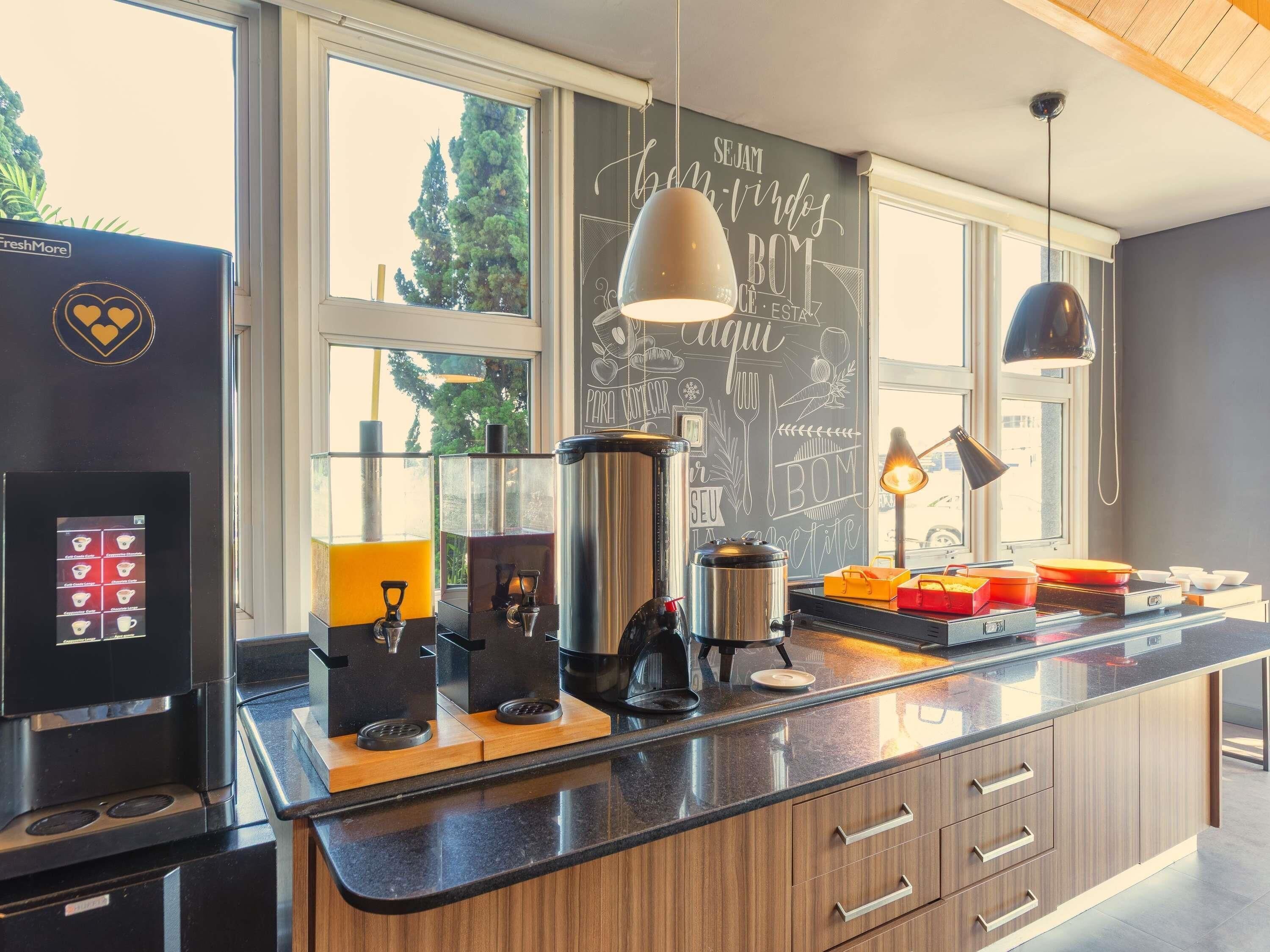 Une cuisine moderne équipée d’une machine à café, d’une bouilloire et d’une cafetière à piston. Des fenêtres offrent une vue sur les arbres, et un tableau noir est accroché au mur.