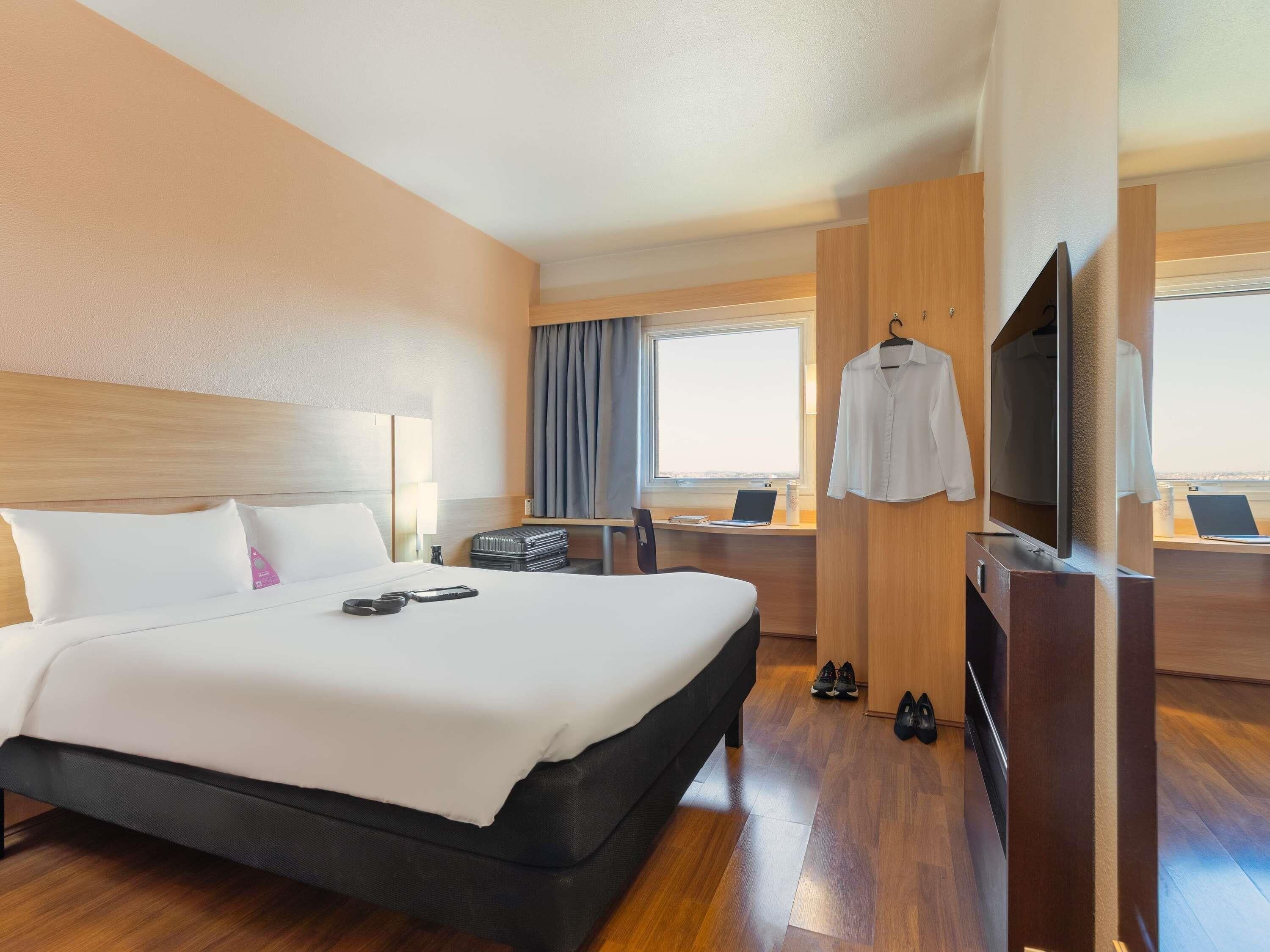 Una habitación de hotel con cama, escritorio con computadora, televisor y ventana con cortinas.