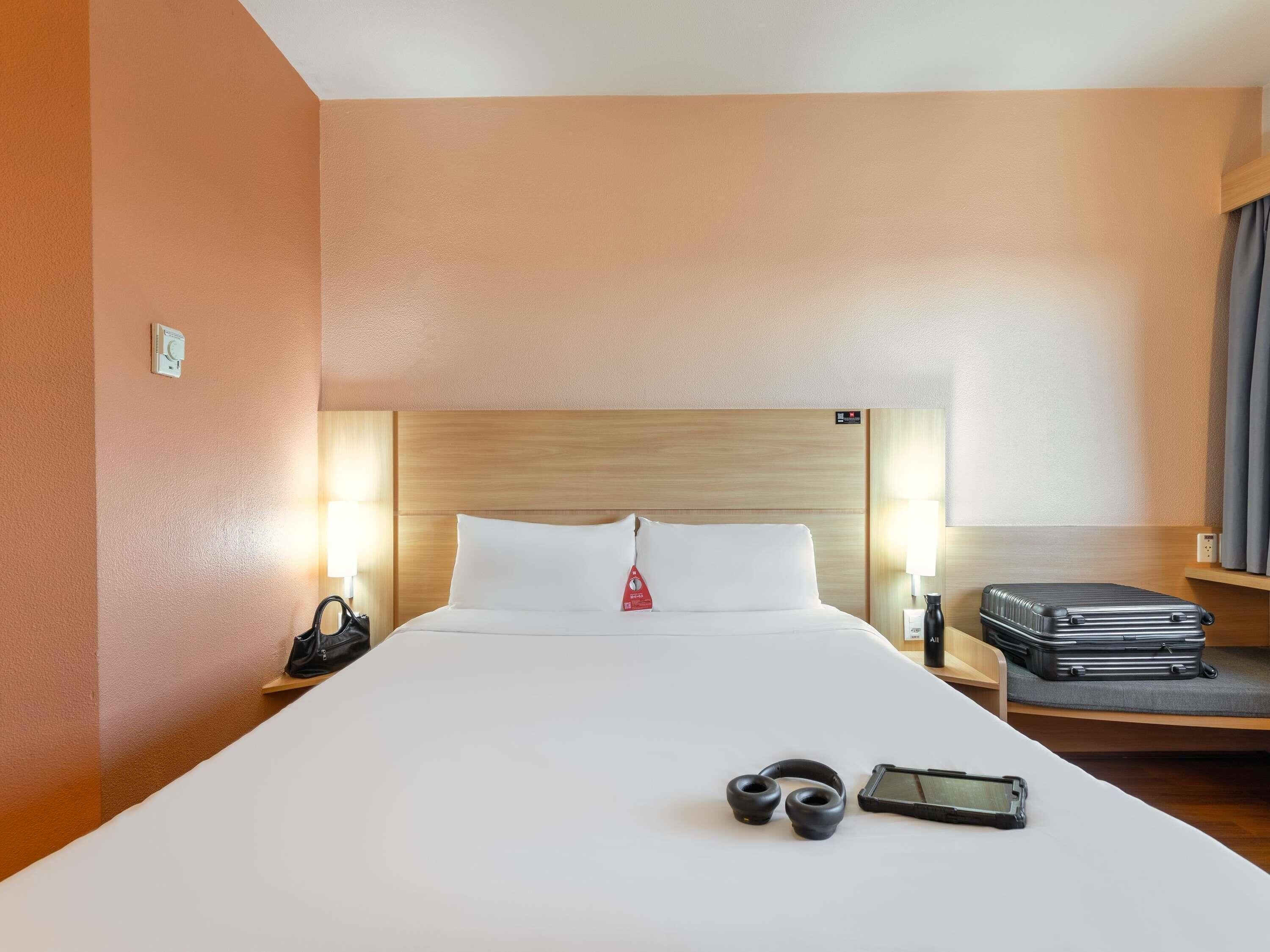 Une chambre d’hôtel avec un grand lit, des lampes de chevet, une valise et une paire d’écouteurs.