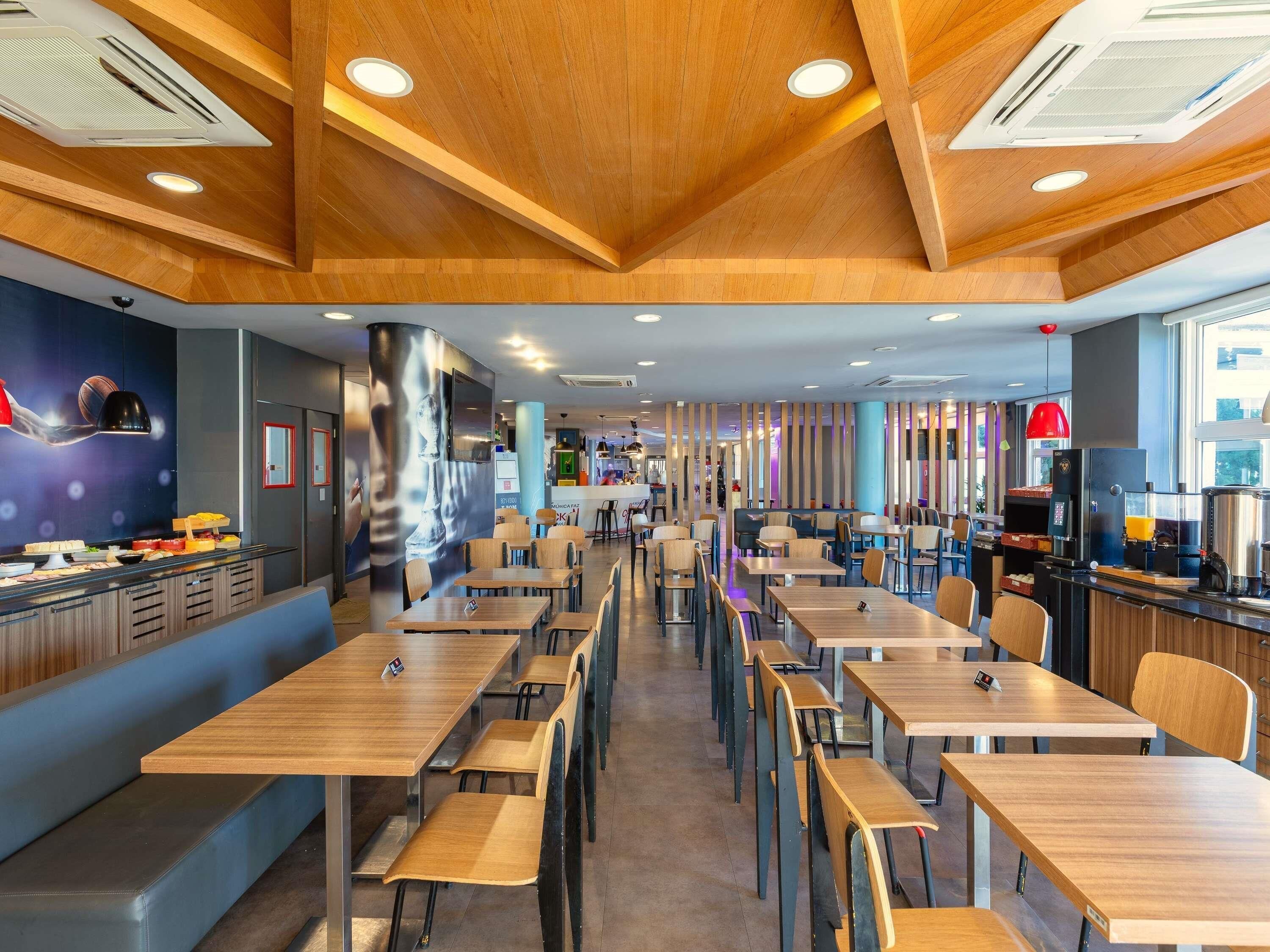 A modern restaurant with wooden ceiling beams, multiple dining tables, and a bar area.