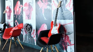 Une pièce dont un grand mur est décoré d’une fresque représentant les membres du groupe, avec deux chaises rouges et une guitare électrique blanche.