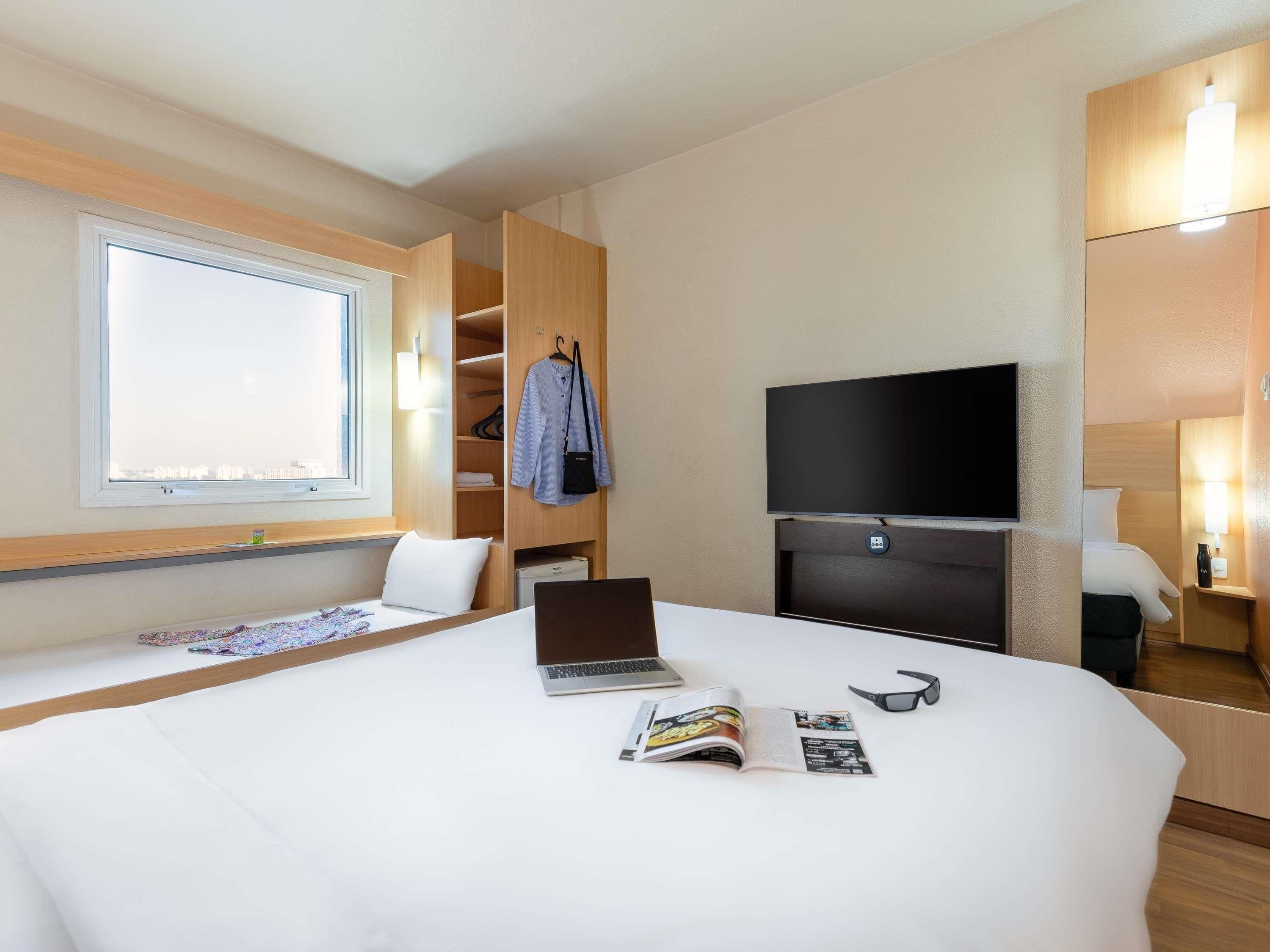 Hotellihuone, jossa on sänky, kannettava tietokone, lehti, aurinkolasit, televisio ja ikkuna, josta avautuu näkymä kaupunkiin.