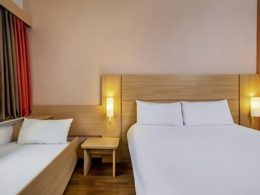 Quarto de hotel com duas camas, uma mesa pequena e duas luminárias fixadas na parede.