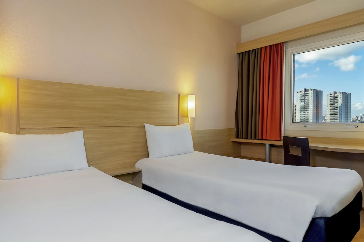A hotel room with two beds, a desk, a chair, a window with a city view, and two wall-mounted lamps.