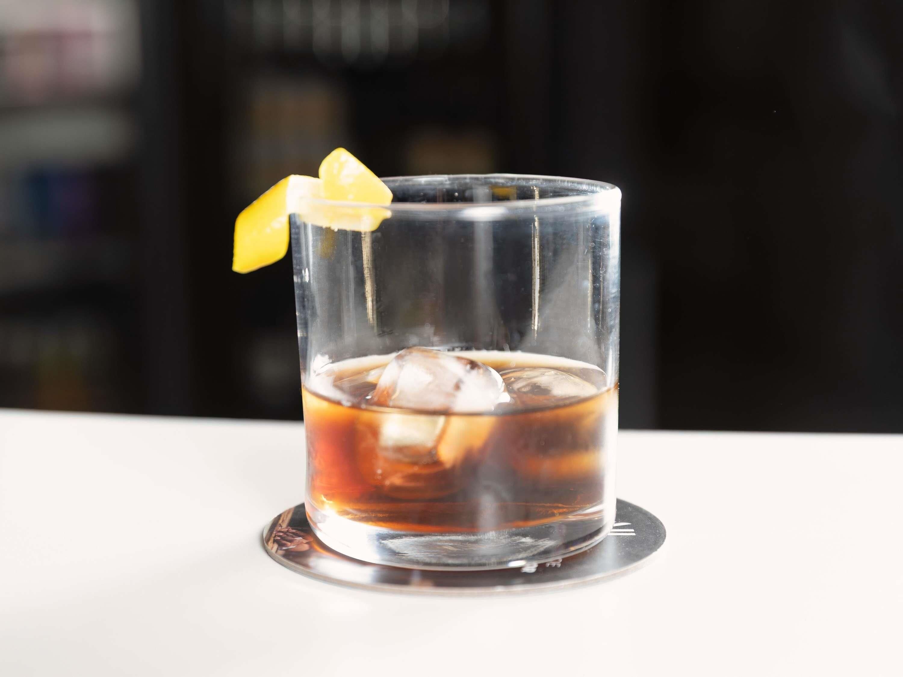Un vaso transparente con un líquido oscuro, cubitos de hielo y una rodaja de limón sobre un posavasos.