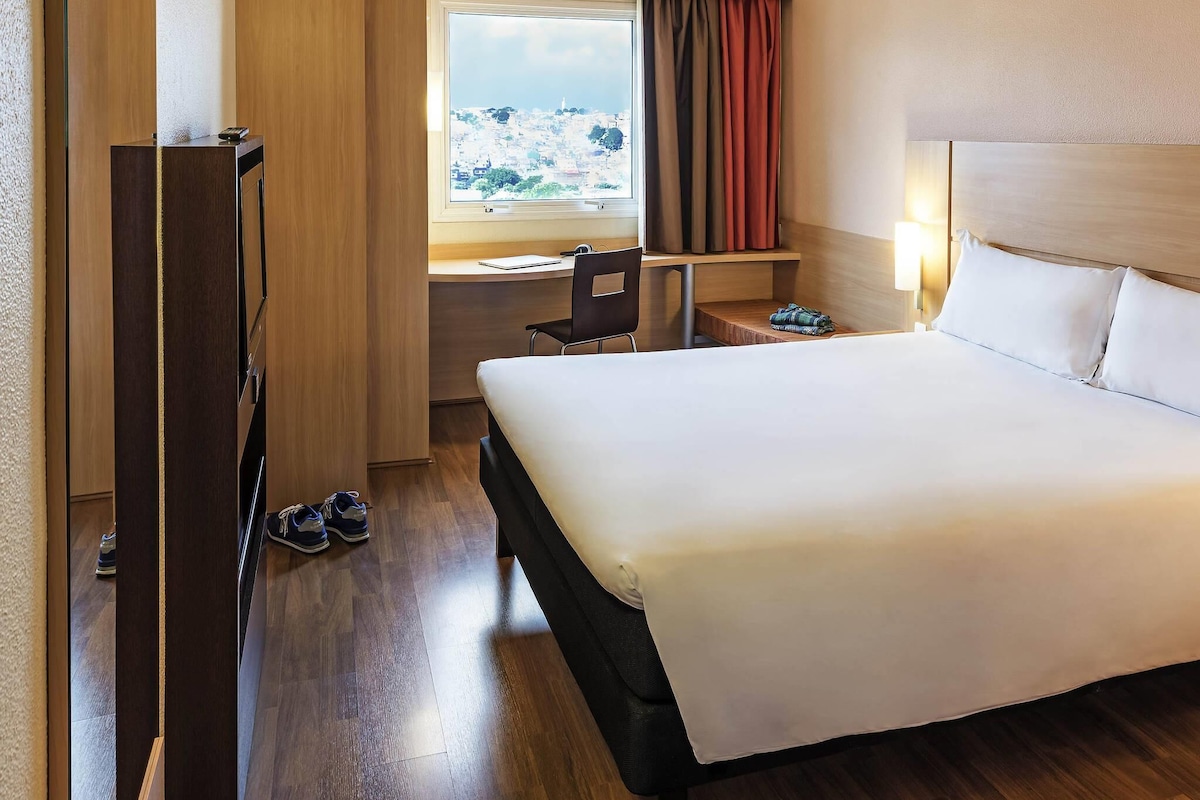 A hotel room with a bed, a desk, a chair, a window with curtains, and a view of a cityscape.