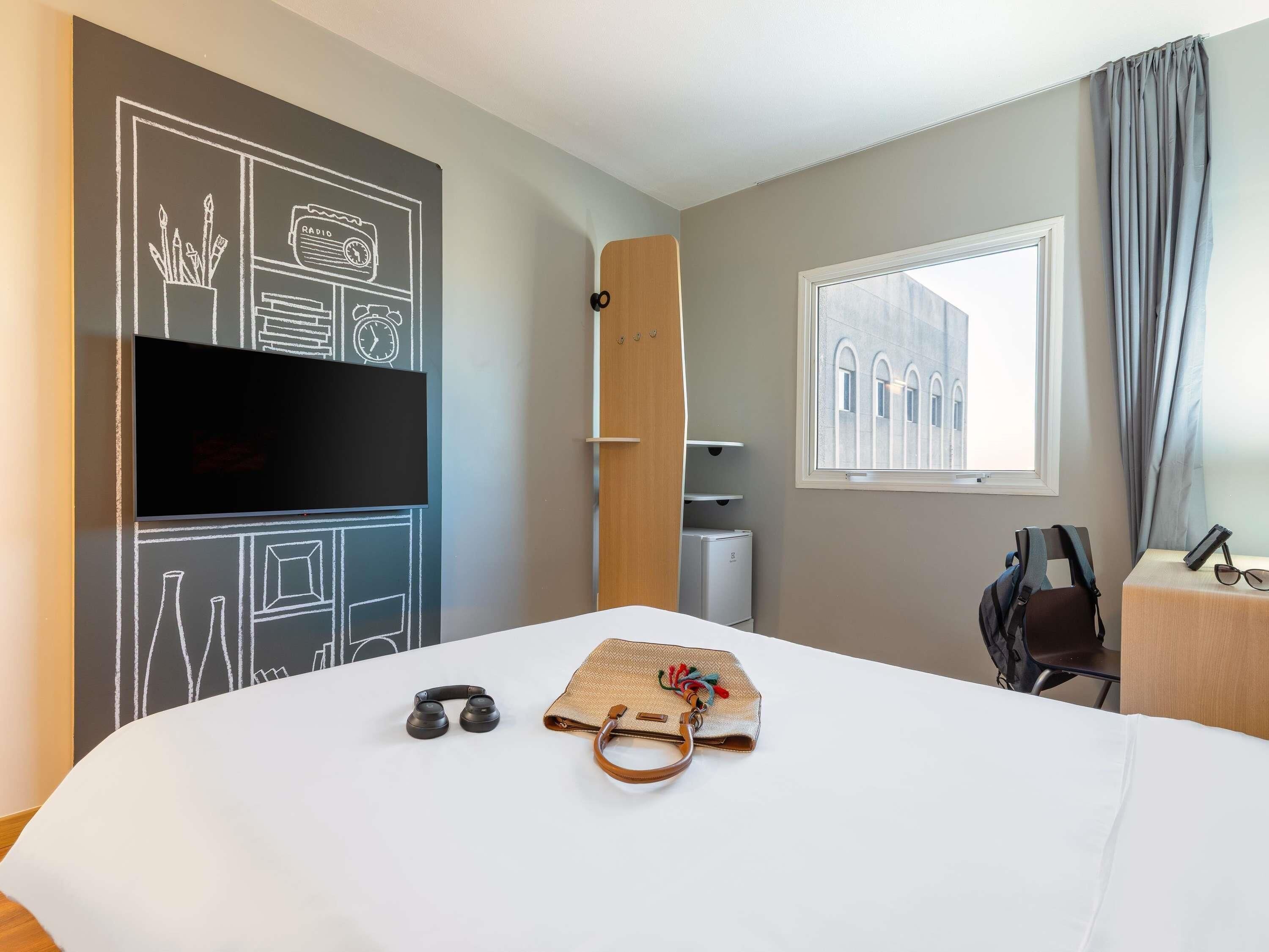 A hotel room with a bed, a TV, a window with a view of a building, a desk with a chair, and a wall with a chalkboard drawing.