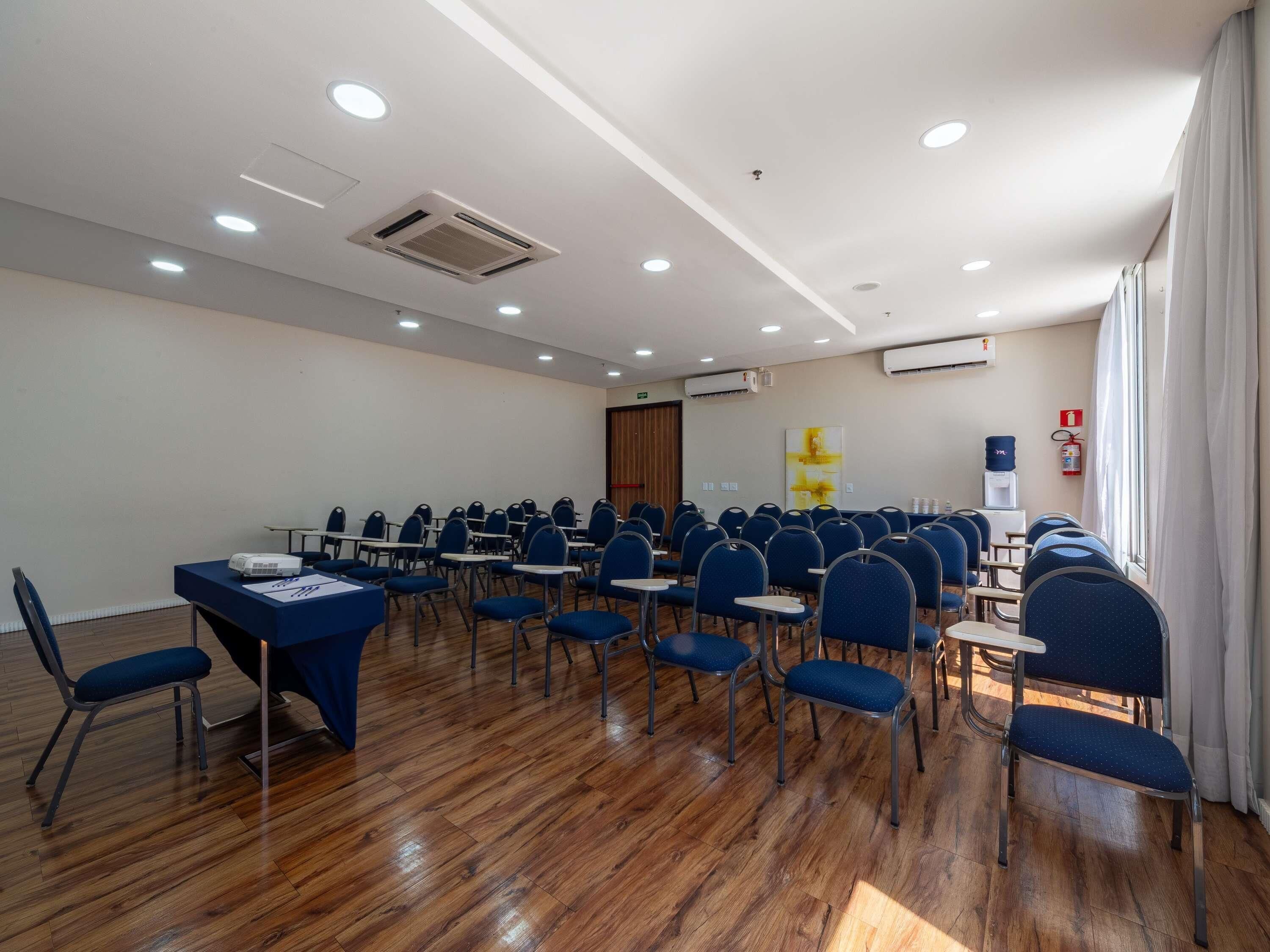 A conference room with rows of chairs, tables, ceiling lights, and air conditioning units.