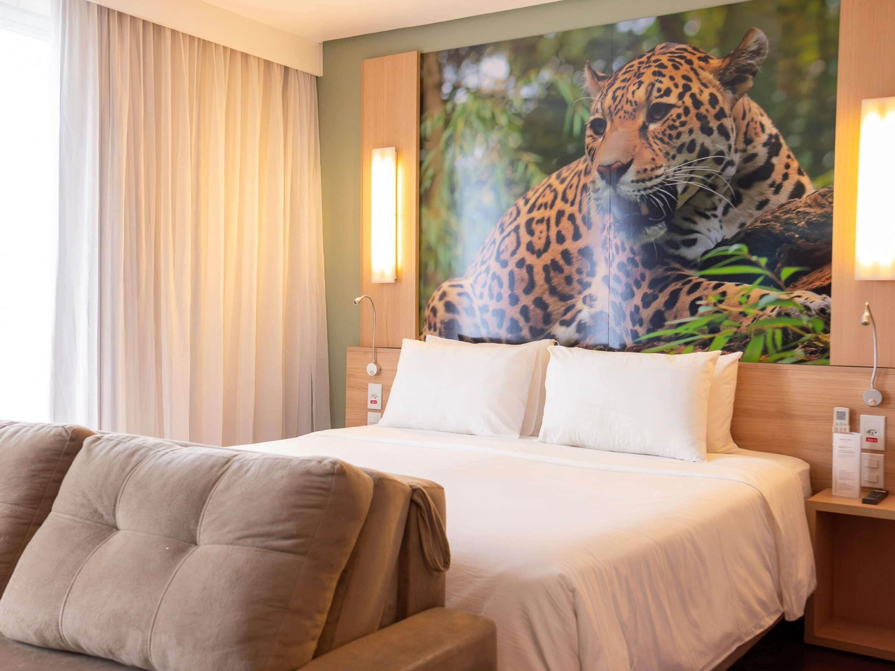 Una habitación de hotel con cama, sofá, un gran mural de leopardo y una ventana con cortinas.