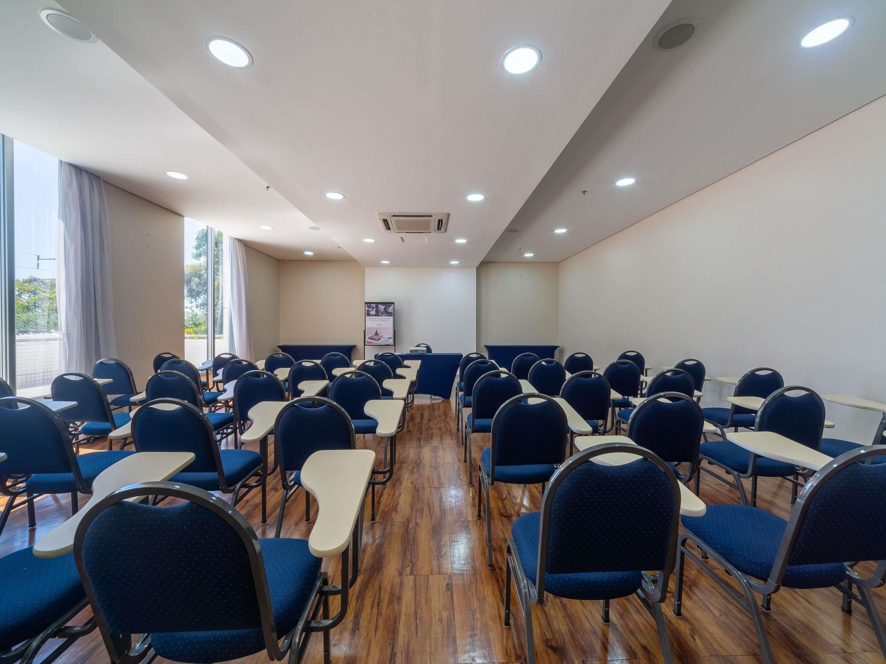 Un salón de conferencias con filas de sillas y mesas azules, un atril y un gran ventanal con cortinas.