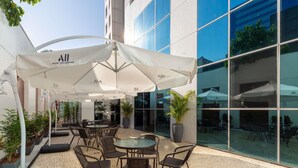 An outdoor patio area with tables and chairs under white umbrellas, surrounded by potted plants and a building with large windows. - Mercure Manaus Hotel (Manaus)
