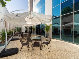 Un área de patio al aire libre con mesas y sillas bajo sombrillas blancas, rodeada de plantas en macetas y un edificio con grandes ventanales.