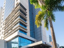Un hotel moderno con fachada de vidrio, una palmera en primer plano y un cielo azul despejado.
