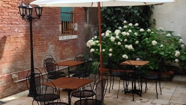 Een terras buiten met betegelde vloer, metalen stoelen en tafels onder een witte parasol.