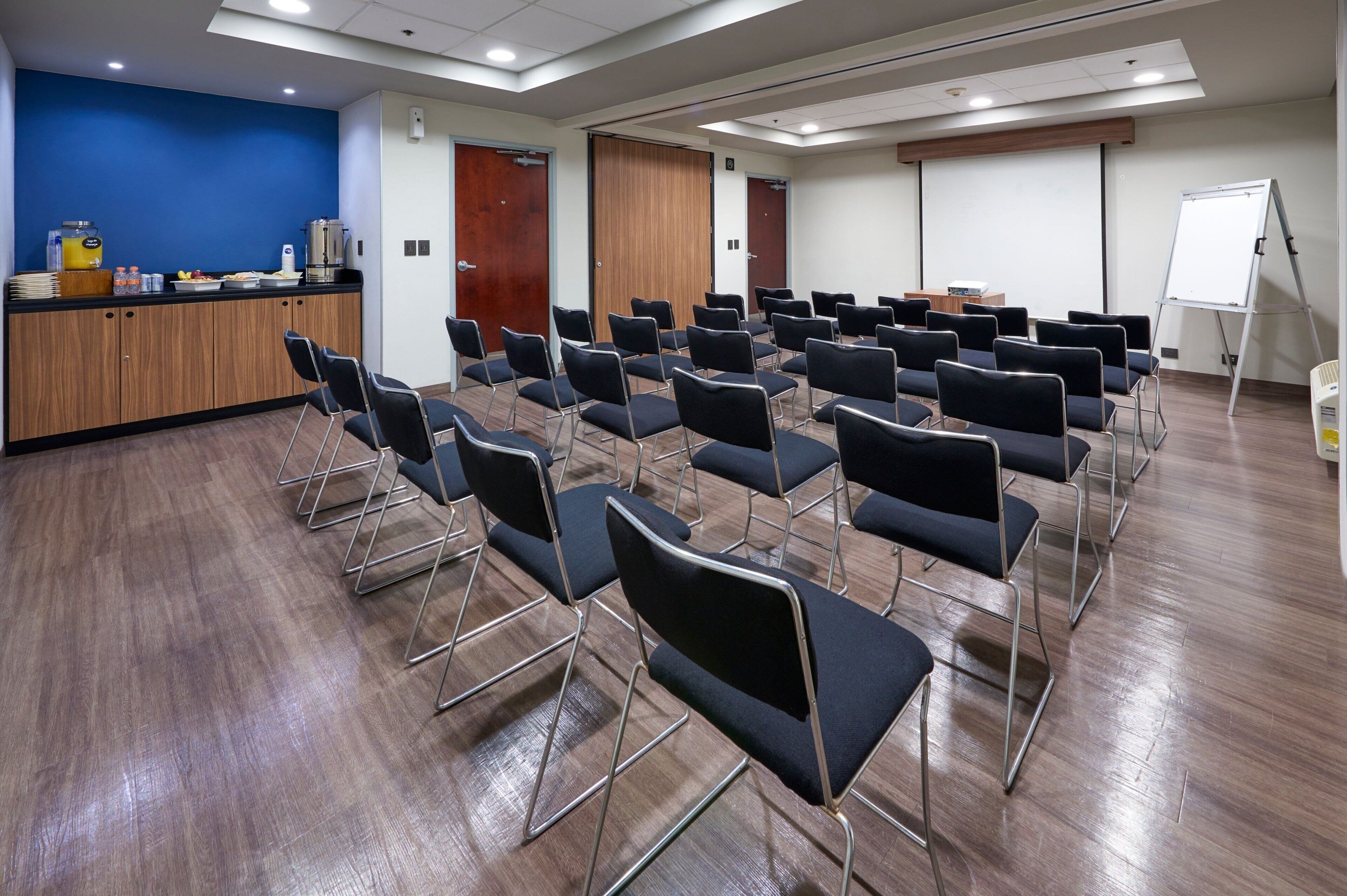 A conference room with rows of chairs, a podium, a whiteboard, and a kitchenette area with food and beverages.