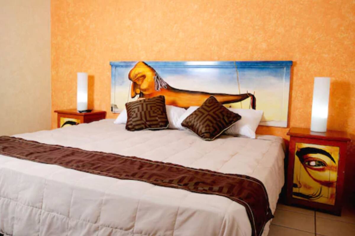 Un lit avec une couvre-lit à motifs, deux oreillers et une tête de lit ornée d’un tableau. Il y a deux tables de chevet avec des lampes.