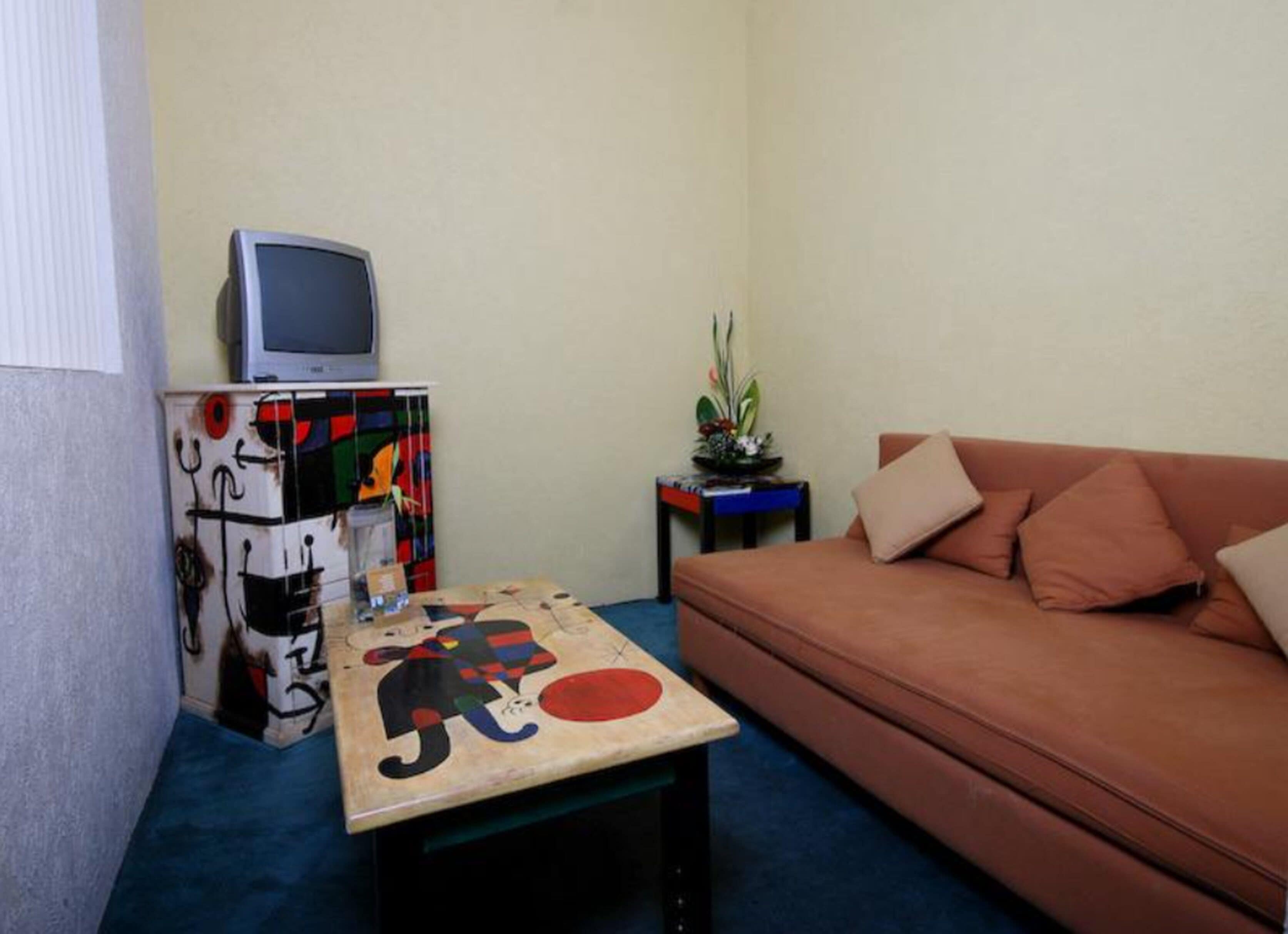 Un petit salon avec un canapé, un téléviseur posé sur un meuble, une table au motif coloré et une plante.