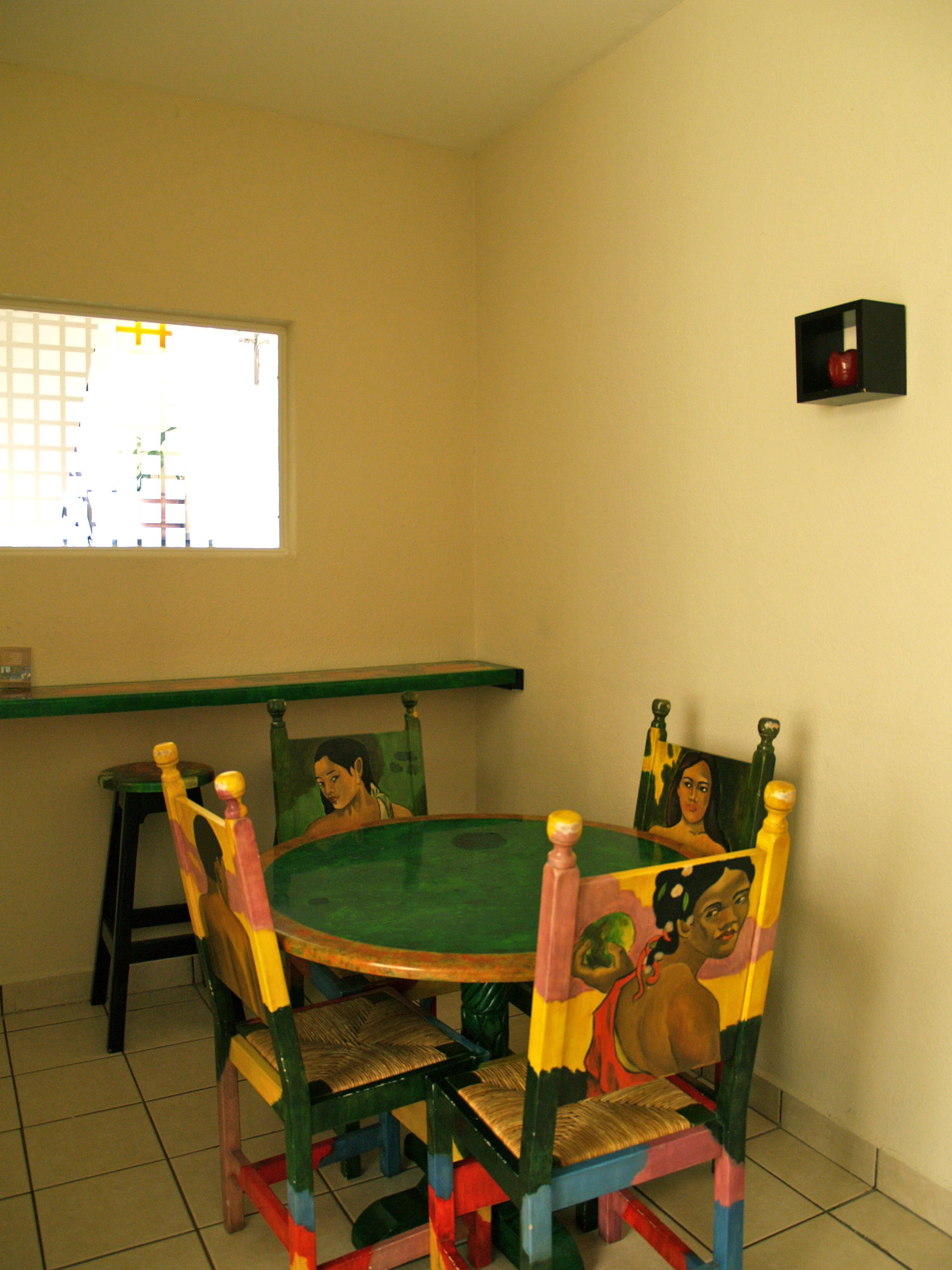 Un comedor con una mesa verde y sillas de colores, una ventana con vistas, y un objeto negro pequeño en la pared.