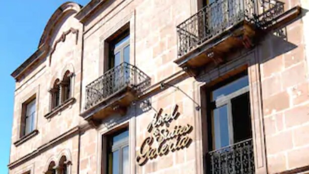 A historic building with a sign reading "Hotel de la Caleta." It features arched windows, balconies with wrought iron railings, and a red awning over the entrance. - Hotel And Suites Galeria (Morelia)