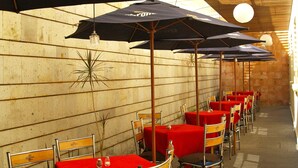 A row of tables with red tablecloths and black umbrellas, arranged in a covered outdoor dining area with a wooden wall backdrop. - Hotel And Suites Galeria (Morelia)