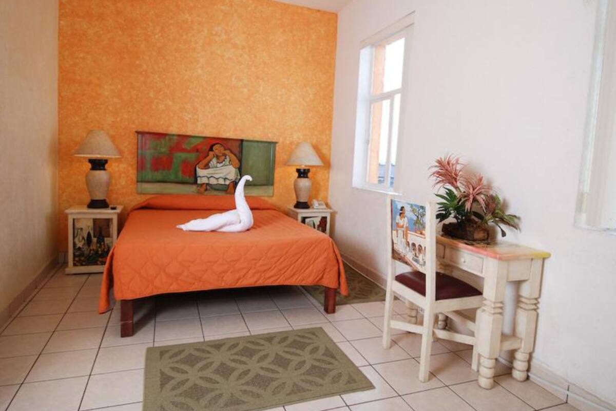 Une chambre avec une couvre-lit orange, deux lampes de chevet, un tableau au mur, une fenêtre et une petite table sur laquelle est posée une plante.