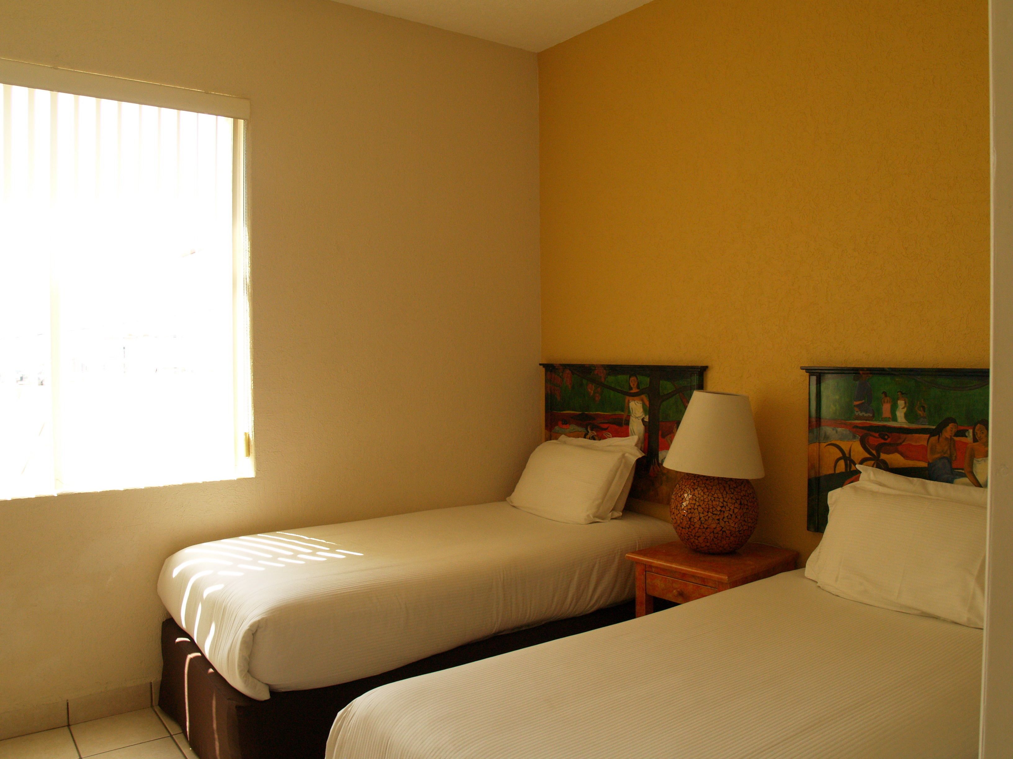 Quarto de hotel com duas camas de solteiro, uma mesa de cabeceira com abajur e um quadro na parede.
