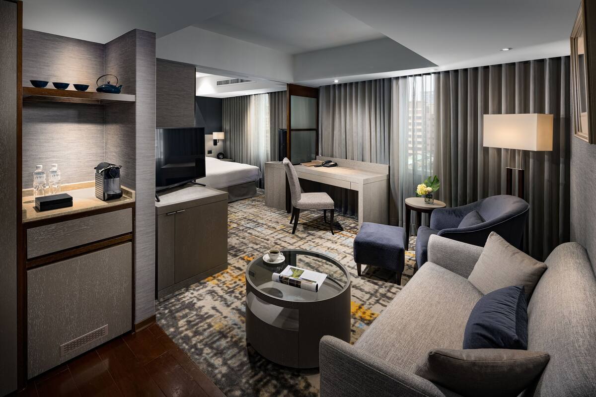 Une chambre d’hôtel moderne équipée d’un canapé, d’une table basse ronde, d’un bureau, d’une télévision et d’un mini-bar.