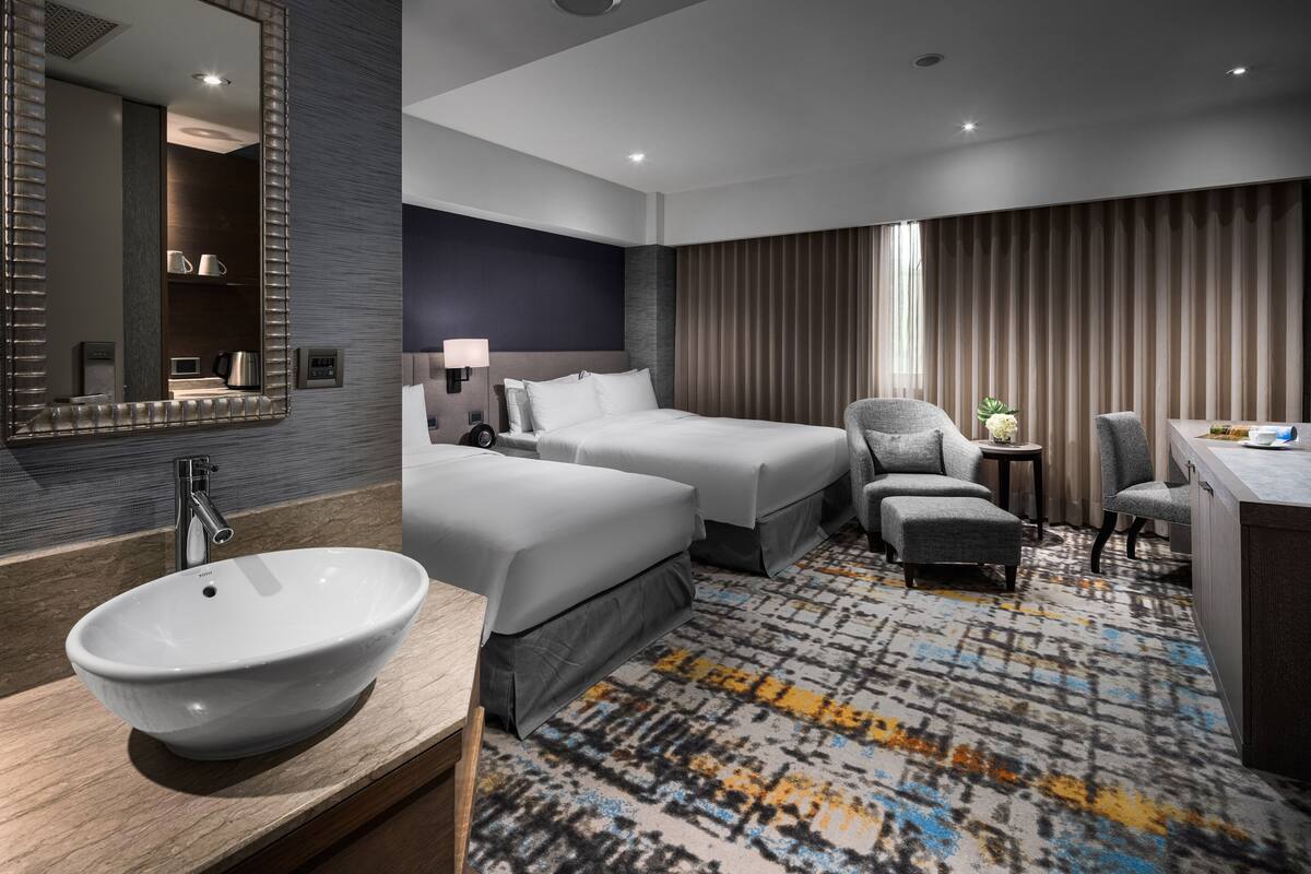 Une chambre d’hôtel moderne dotée d’un grand lit, d’un coin salon, d’un bureau et d’une salle de bain avec un grand lavabo et un miroir.