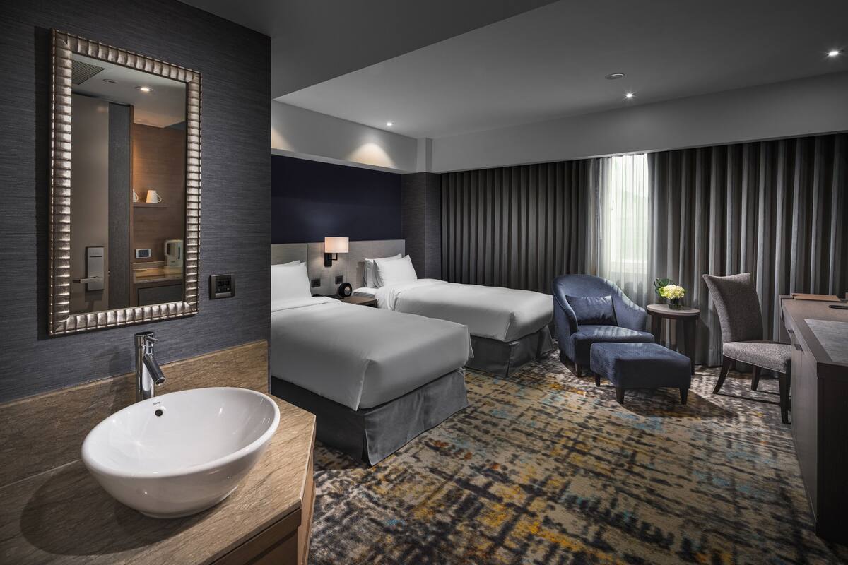 Une chambre d’hôtel avec un grand miroir, un lavabo blanc, deux lits, un bureau et une chaise.